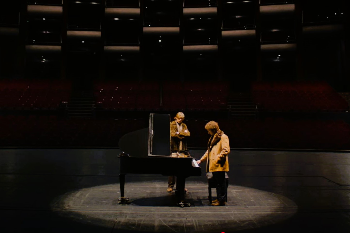 Two people sit and stand next to a grand piano on a stage.