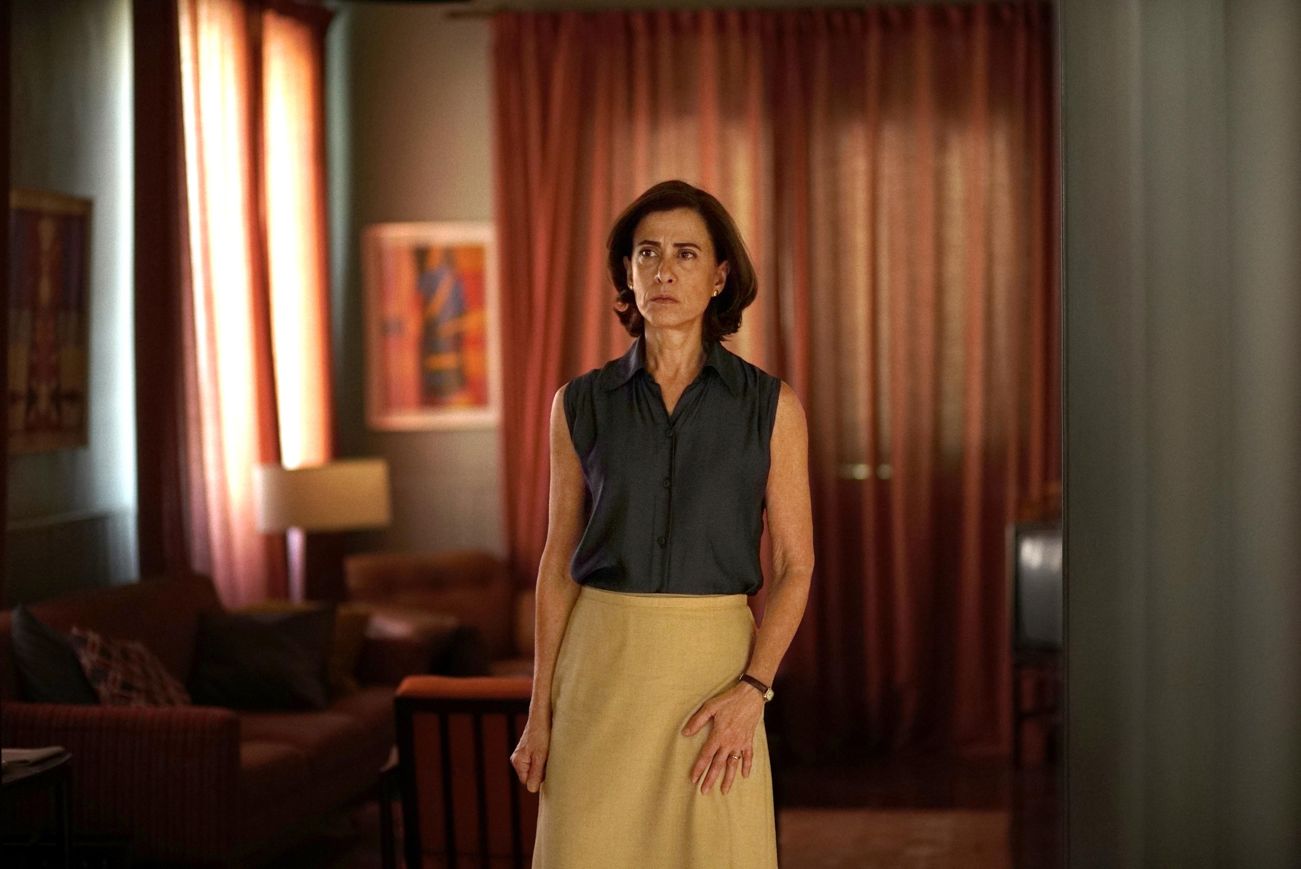 A woman, wearing 70s clothing, stands, looking apprehensive.