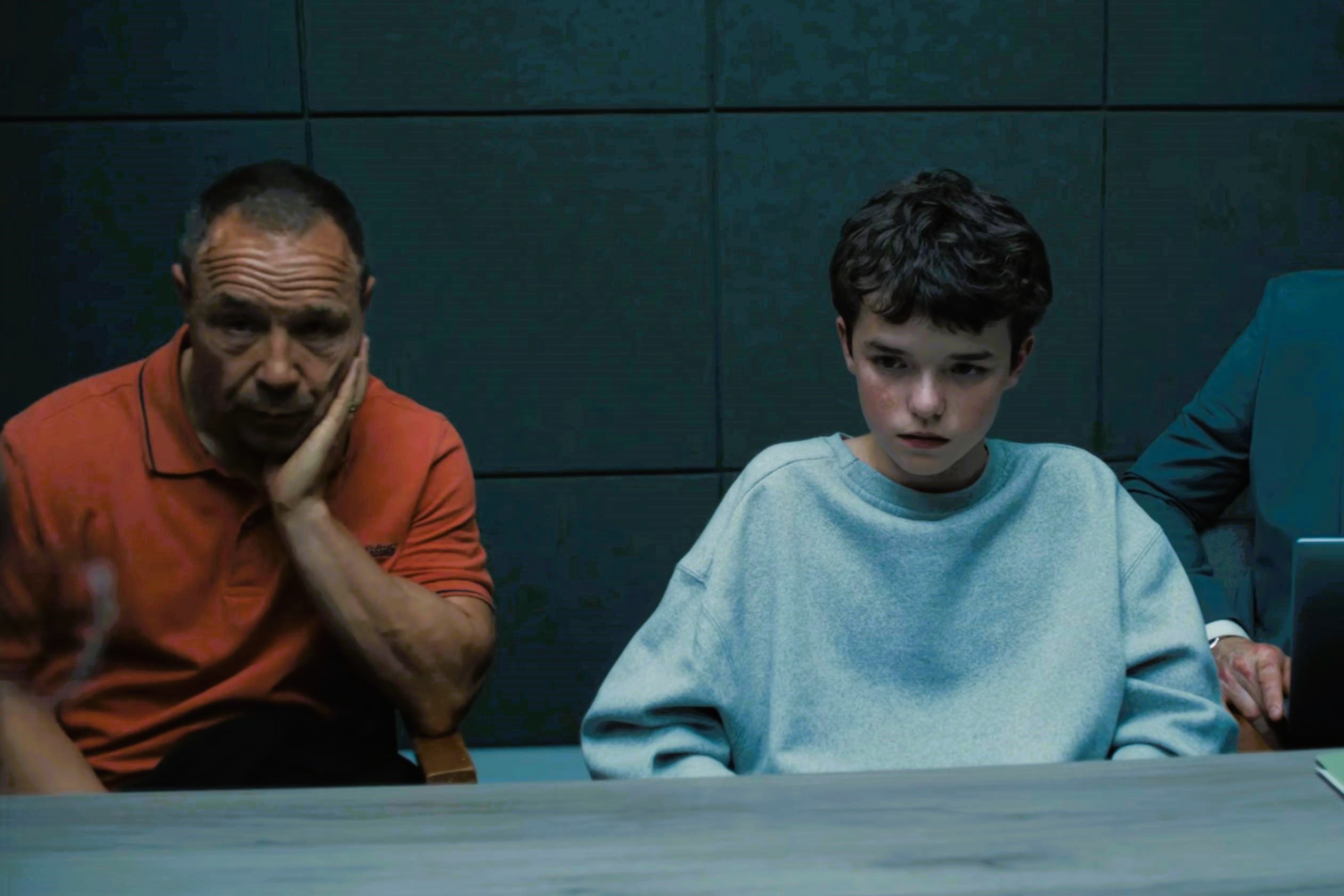 An unhappy father sits next to a scared son in a police interview room.