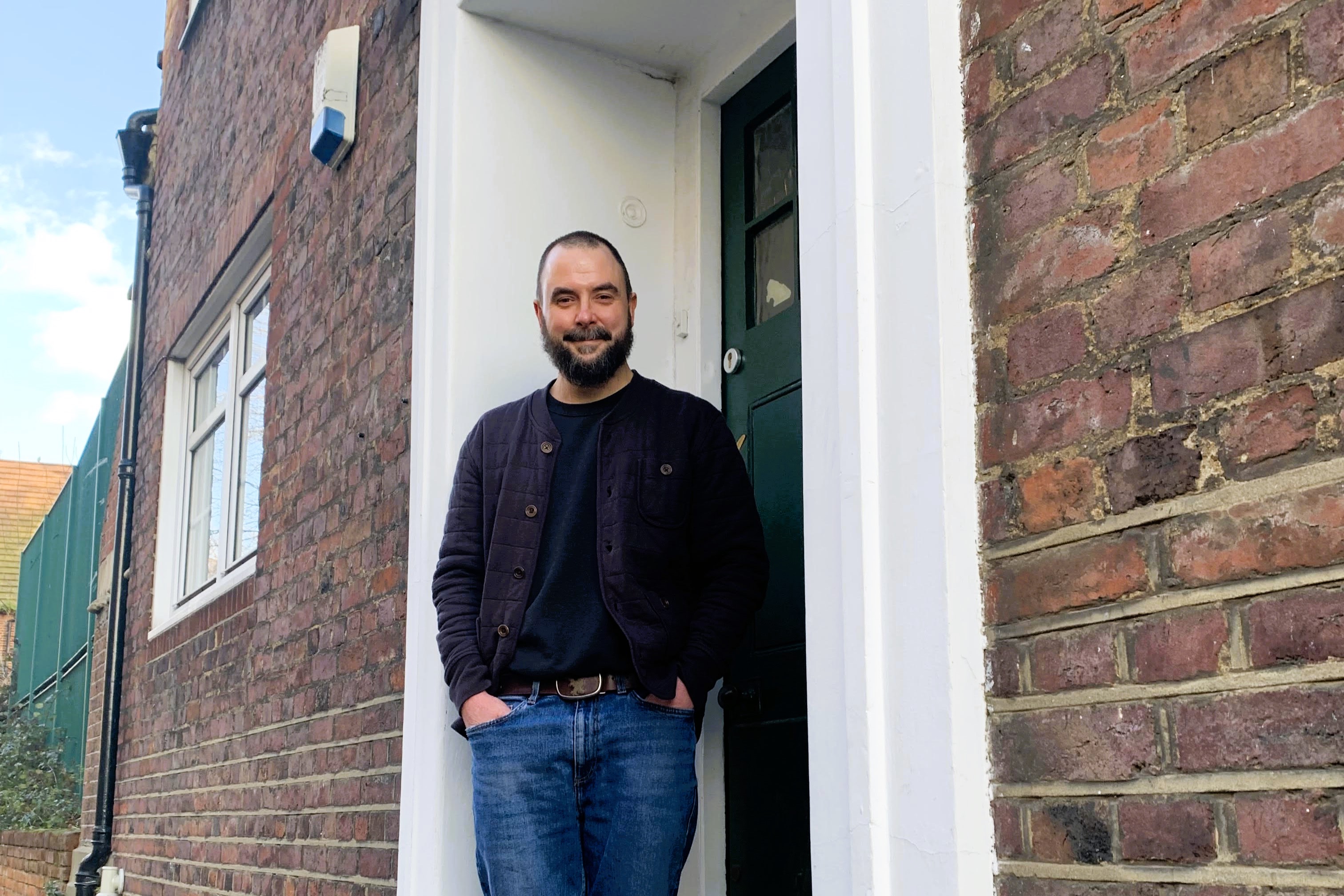 A man stands in a relaxed way on the doorstep of a brick building.