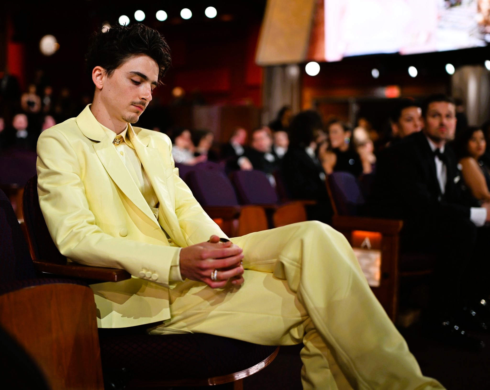 Timothee Chalamet, wearing  light yellow suit reposes in a seat a the Oscars, clasping his hands, his eyes closed.