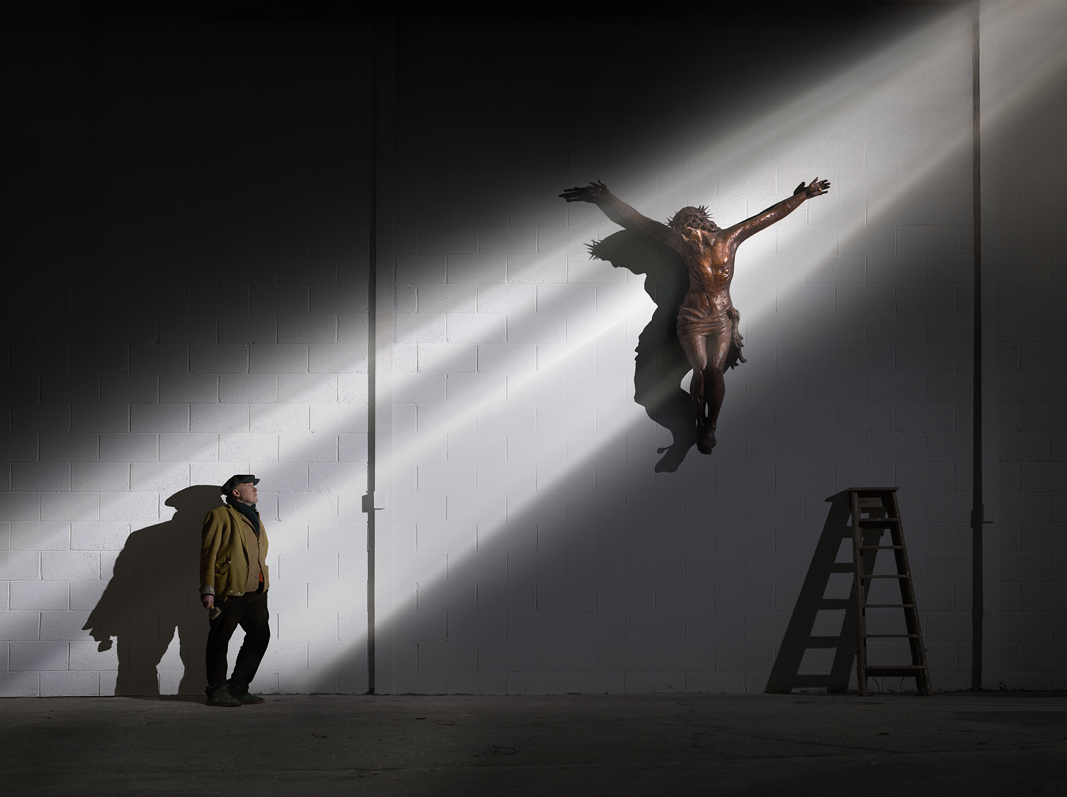 A man looks up a shaft of light that illuminates him and a crucifix higher up a wall.