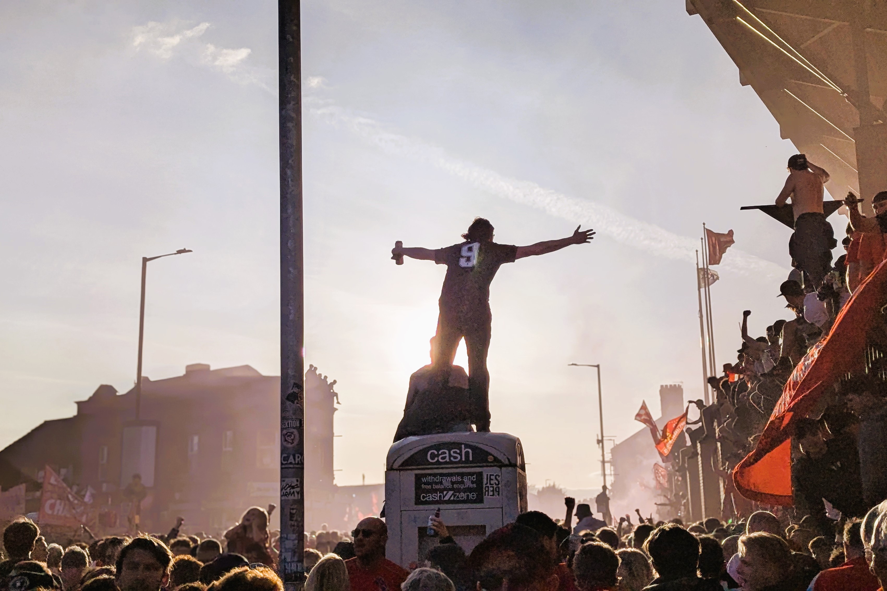 Amid celebrating football fans, one stands on top of a kiosk with outstretched arms.