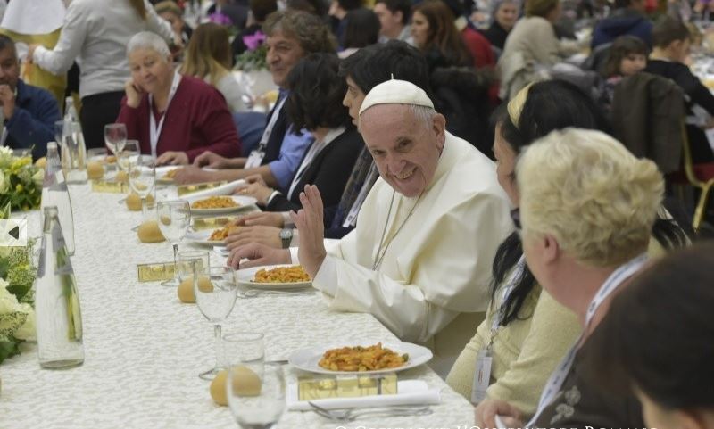 The Pope sits at a table having a meal, raising a hand of blessing