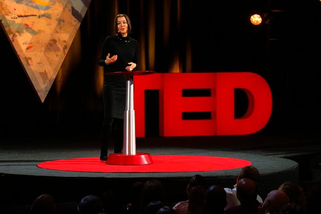 A speaker stands beside a podium in front of a TED sign.
