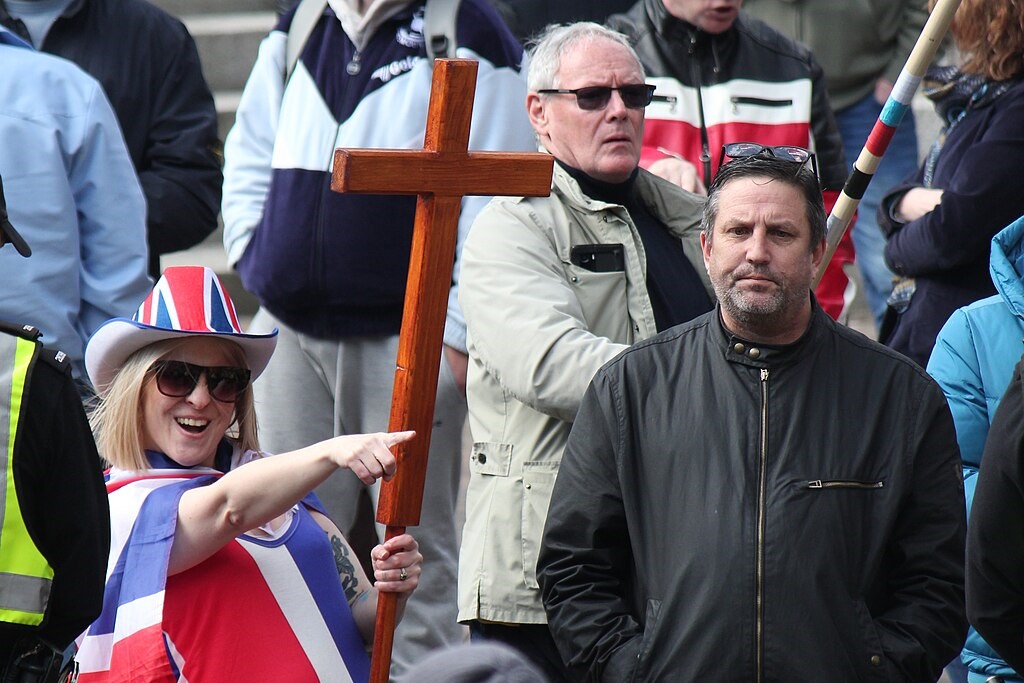 A protester wearing a Union Jack flag and hat and holding a cross, points while a man looks on.