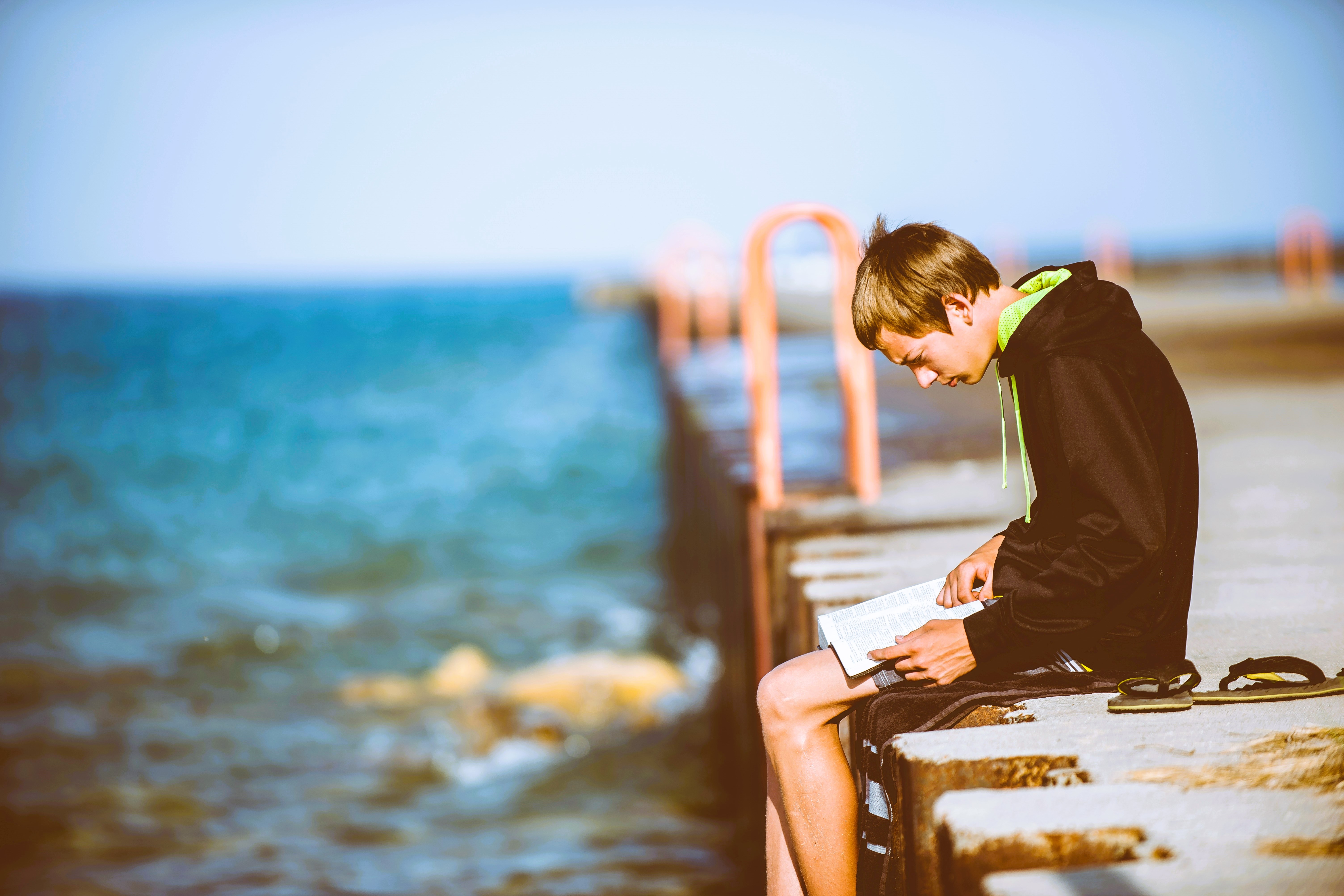 A man sits on a pier intently reading a book on his lap.