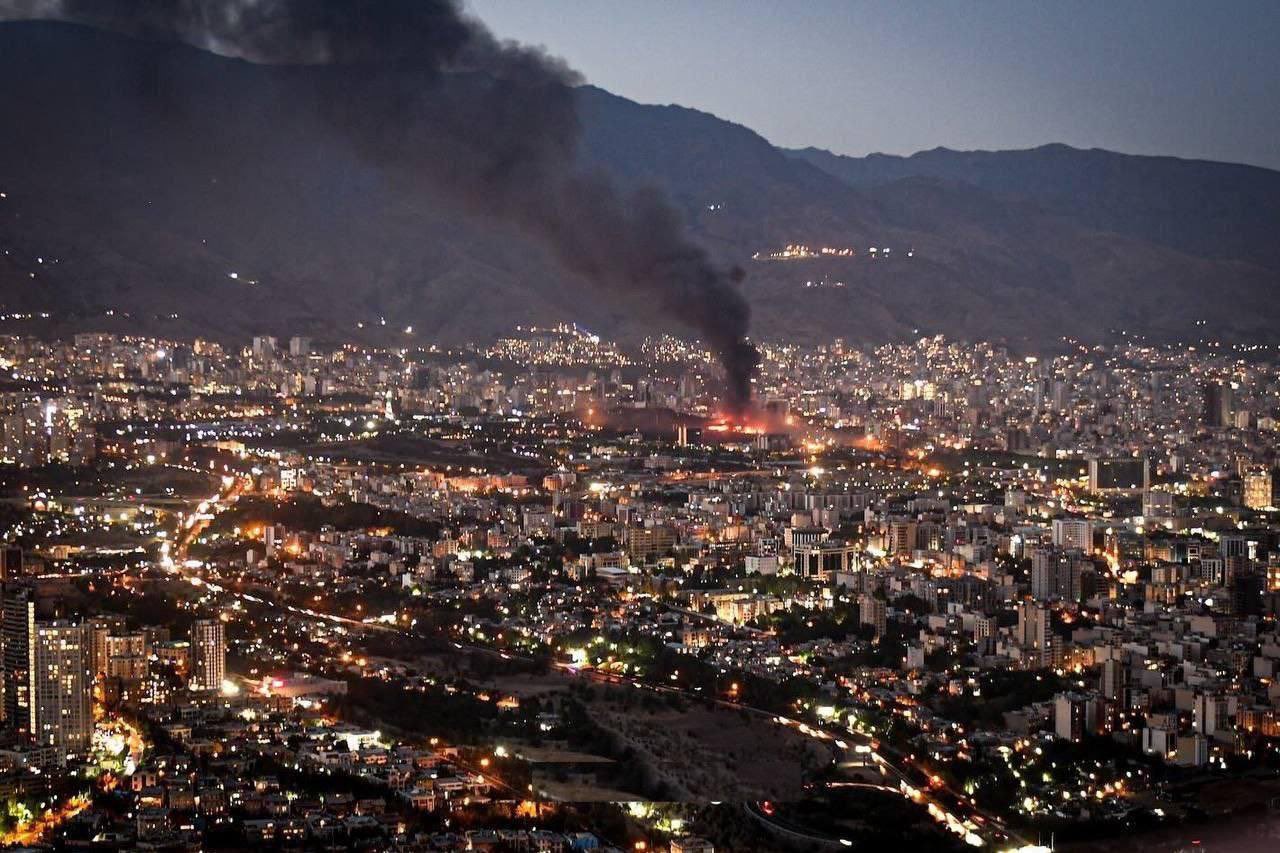 Smoke rises from the site of a bomb amid a high wide view of a city at twlight