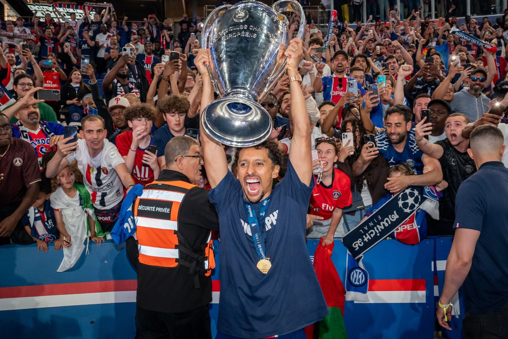 A footballer holds up the European cup, behind him fans are jubillant.