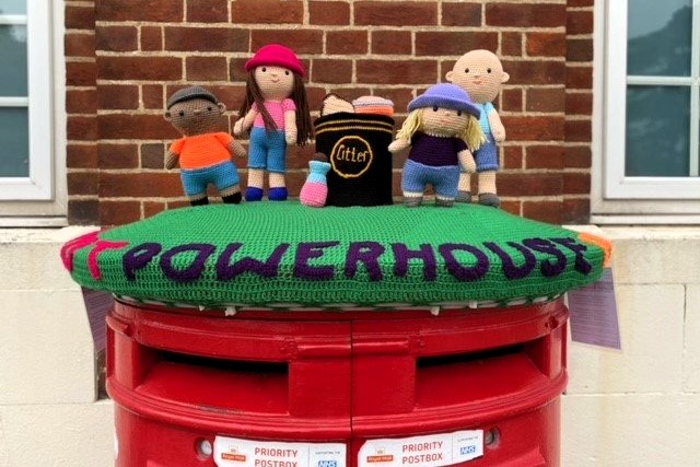 A knitted post box topper shows a group of people and the word powerhouse.