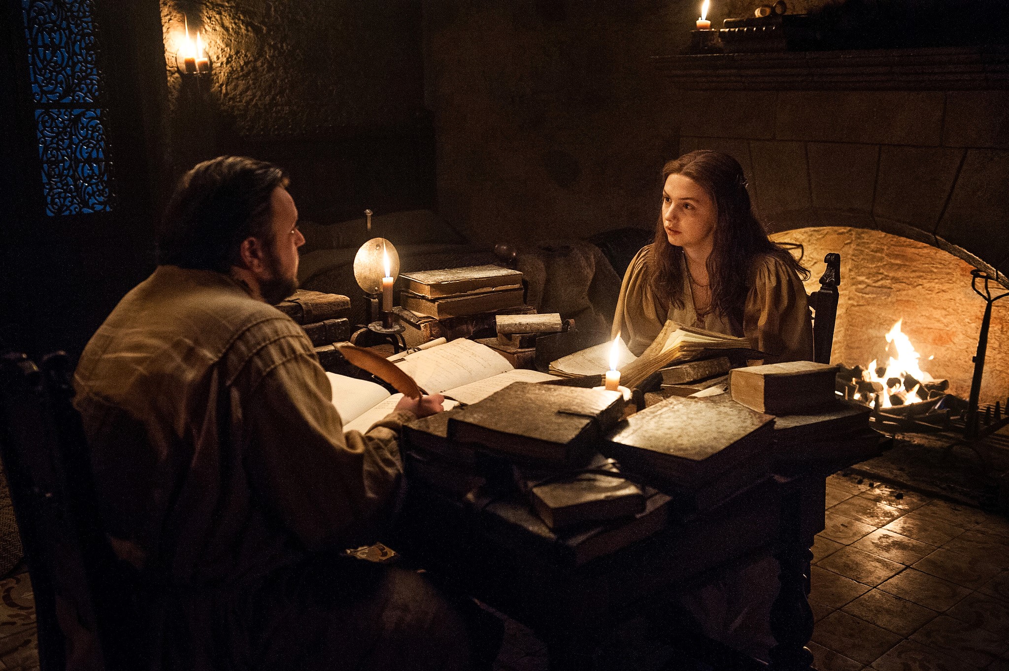Two people sit at a table strewen with old books lit by candle light.