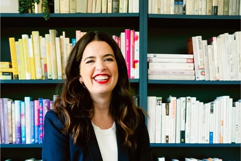 A woman sits and smiles in front of her bookcase.