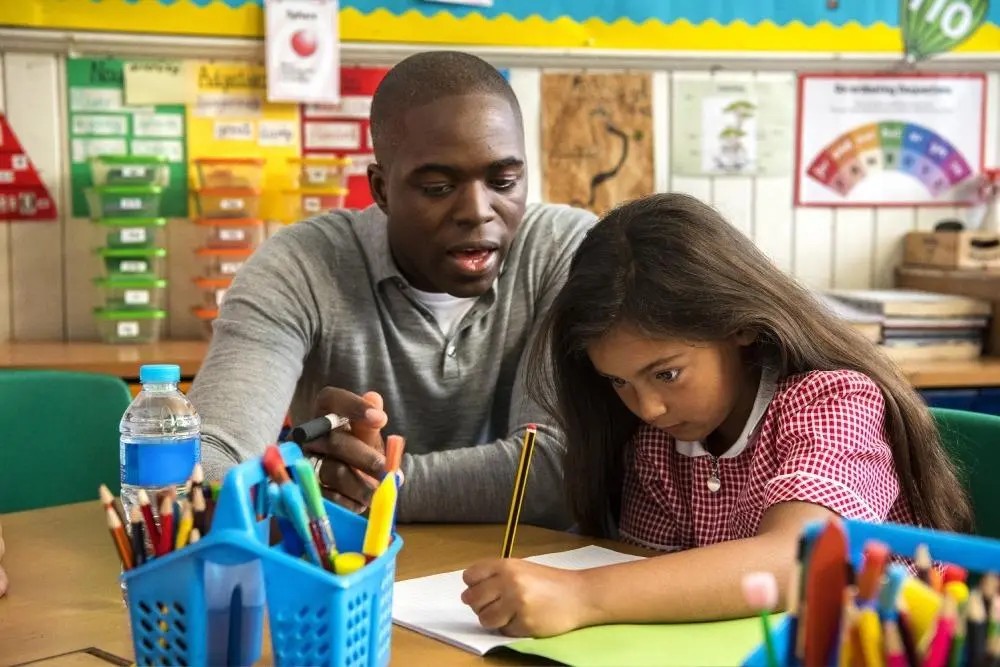 A teacher looks on as a young child concentrates on writing.