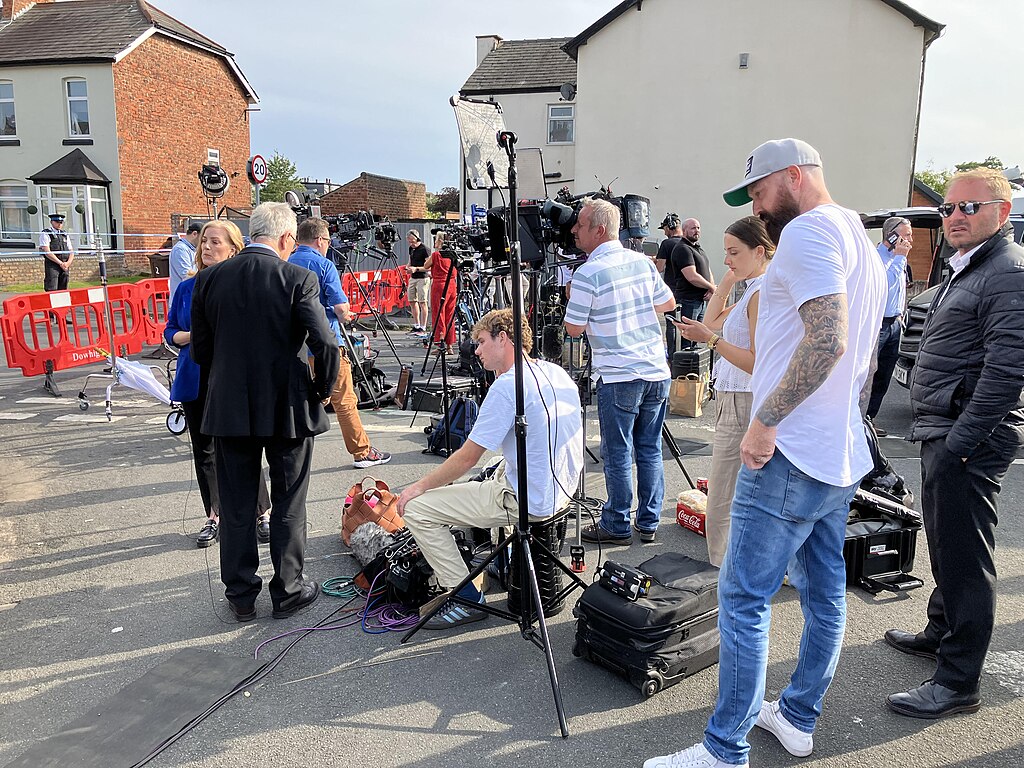 A media pack await a press conference in a street.