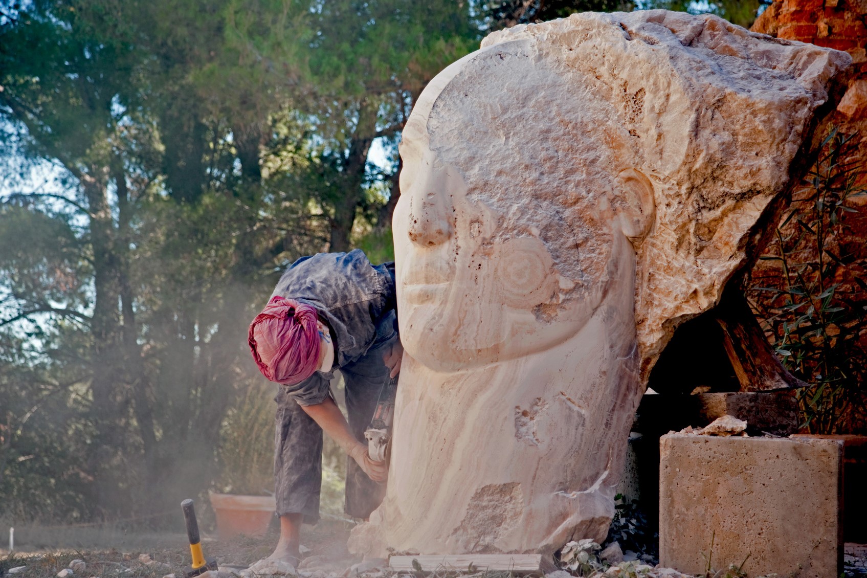 A sculptor works on a carved face emerging from a stone larger than her