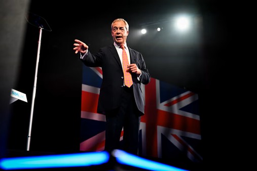 Nigel Farage stands and gestures in front of a flag.