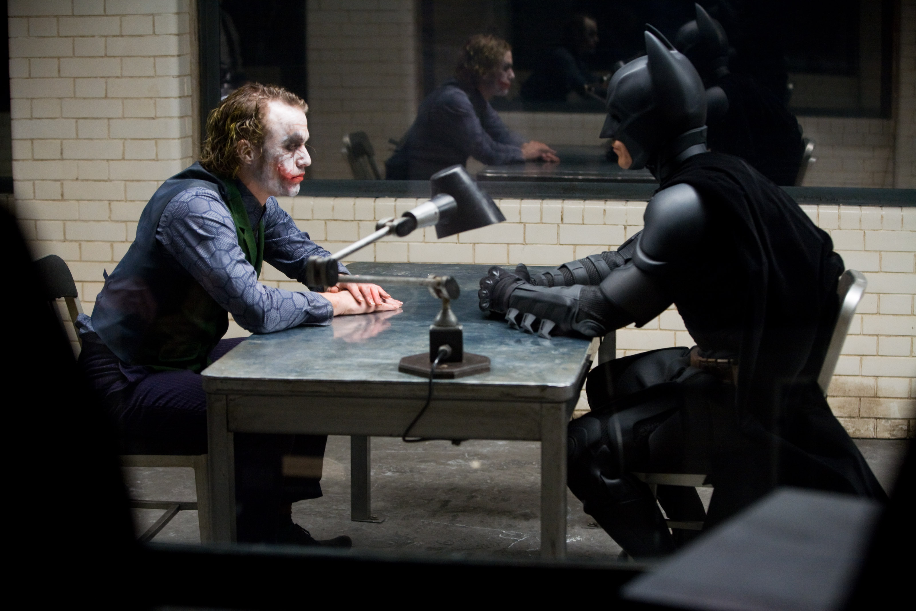 The Joker and Batman sit opposite each other in an interrogation room.