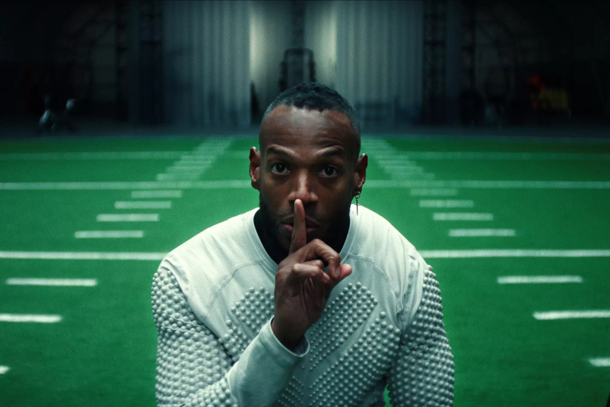 A player holding a finger to his lips stands in front of an indoor American football pitch.