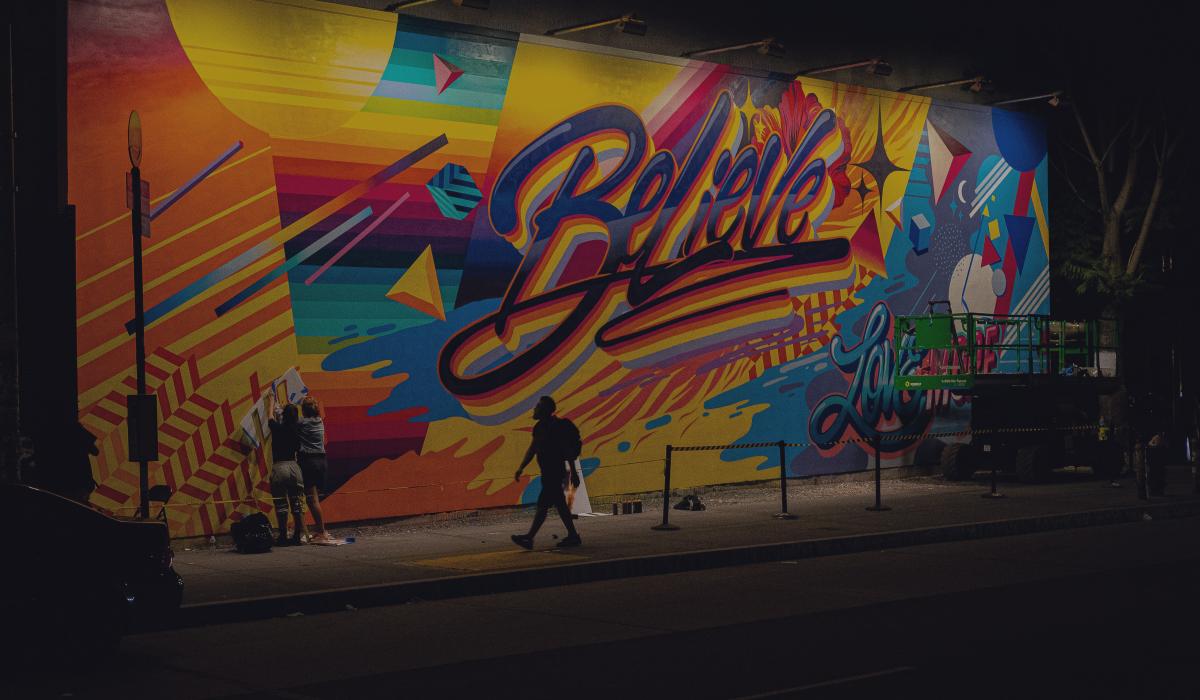 A person walks past a multi-coloured wall of graffiti with the word 'believe' in the centre of it.