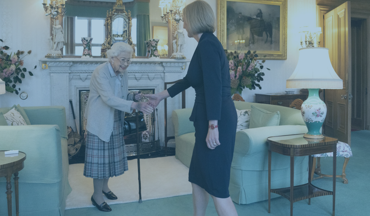 A frail old lady, the late Queen, rises from a sofa to shake hands with an approaching woman.