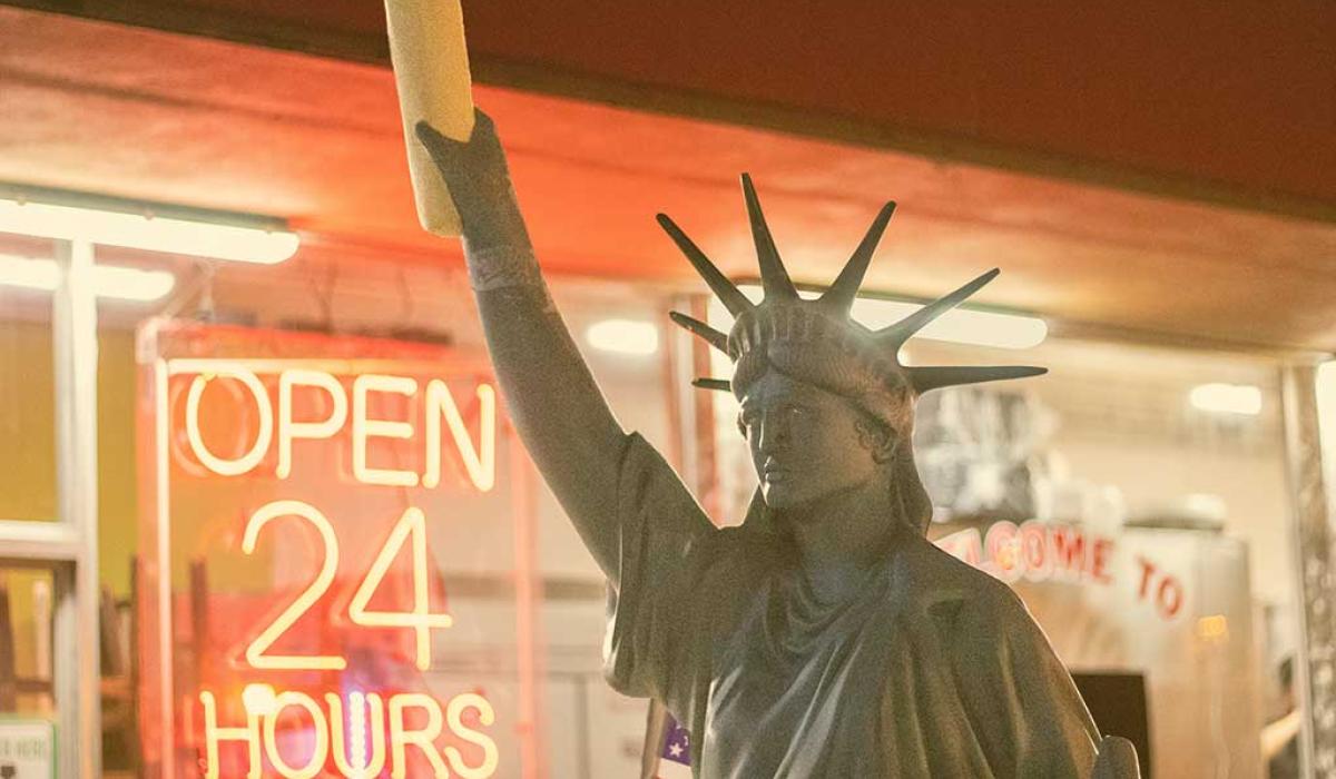 A copy of the Statue of Liberty, holding a stick of bread, stands outside a shop window displaying an 'Open 24 Hours' signs.