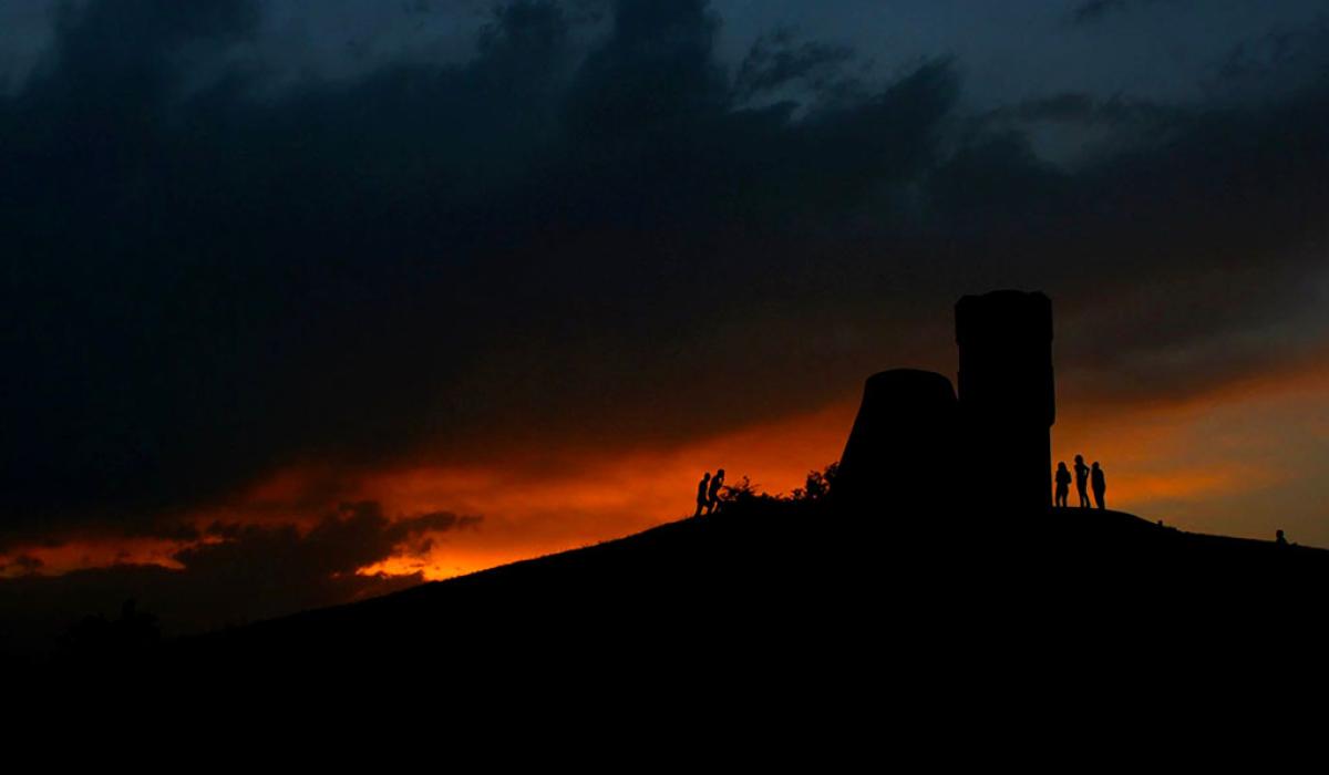 A sunset dramatically silhouette's a ruined tower and people at its base.