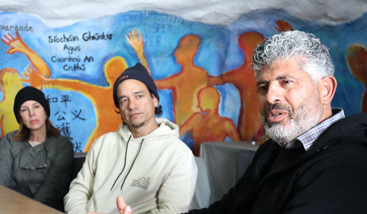 Two people down a table turn and listen to someone closer talk, against a wall mural.