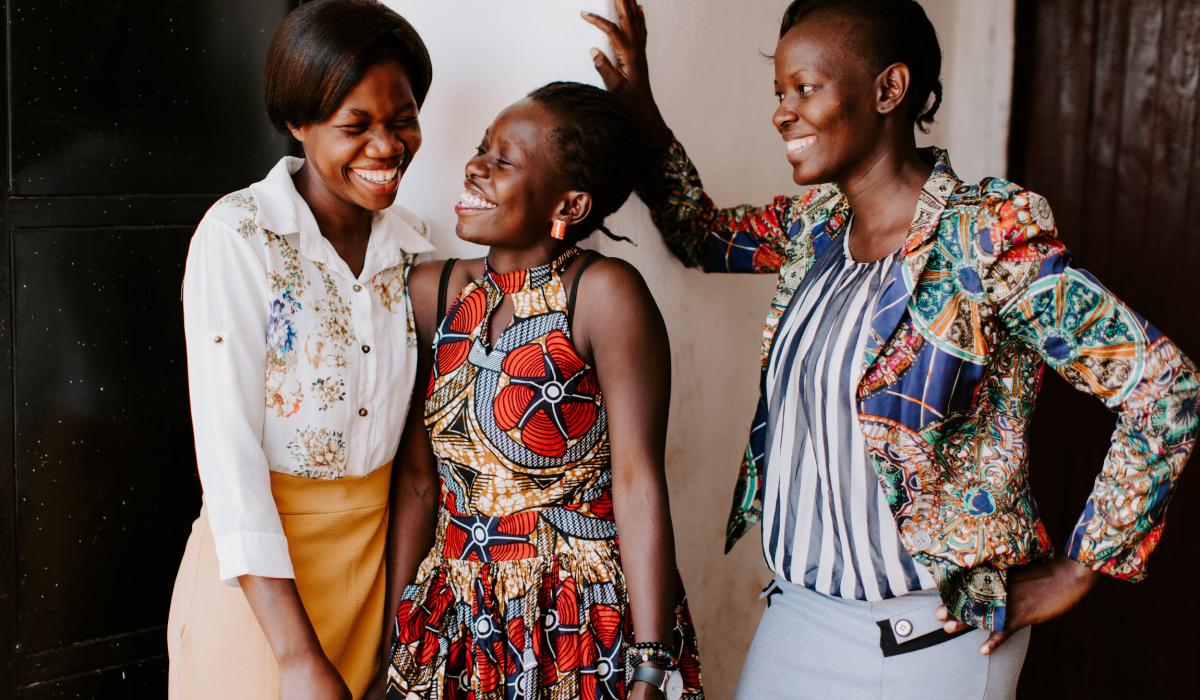 Three women stand, two lean into each other sharing a joke, while the other laughs too.