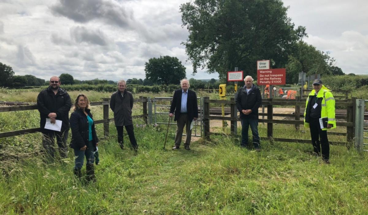 A group of people stand in a field by a fence and a railway footcrossing.