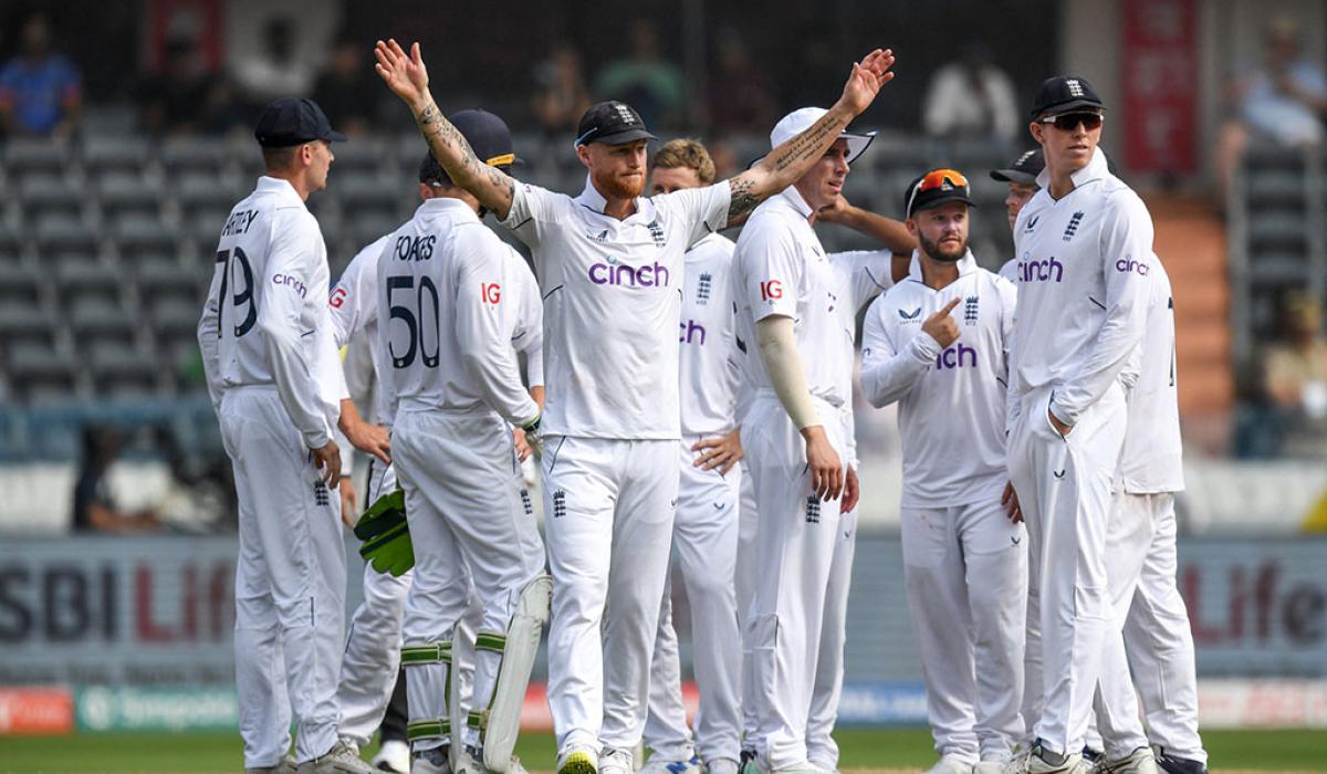 A gaggle of cricket players, dressed in whites, stand on the field. One raises there arm