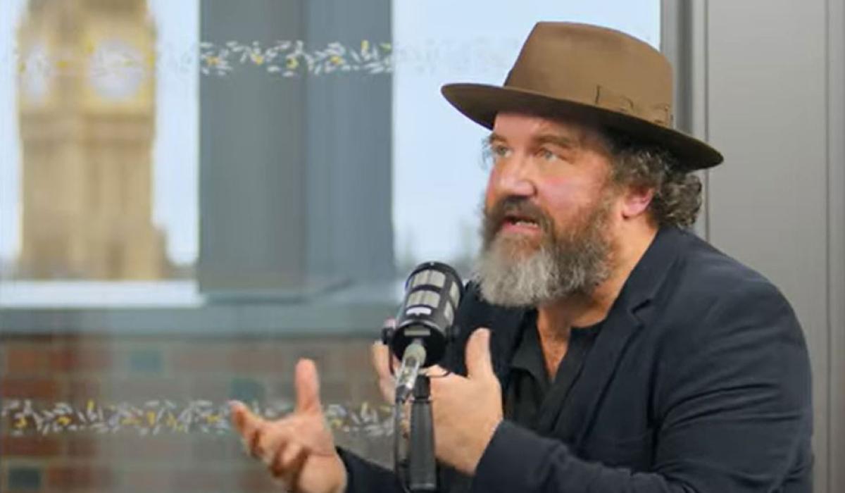 A man wearing a hat sits at a table talking and raises both hands in front of himself to gesture