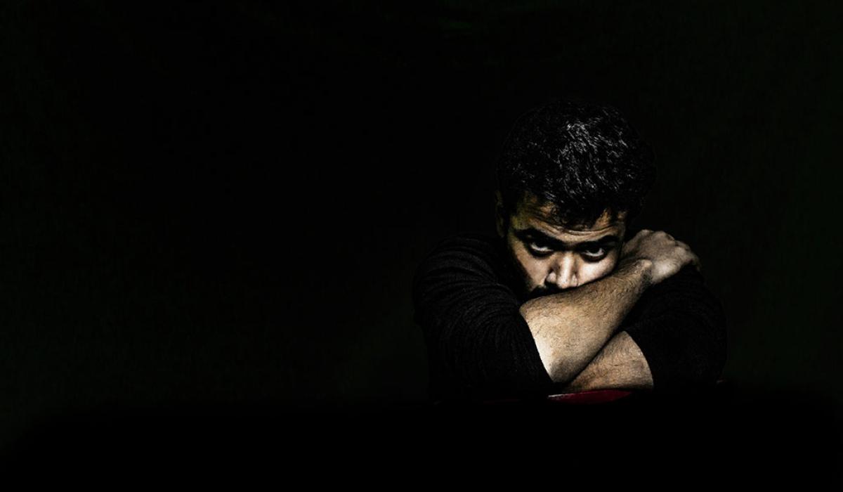 In a darkened room, a man's angry face is lit as he rests on arms folded tightly around it.