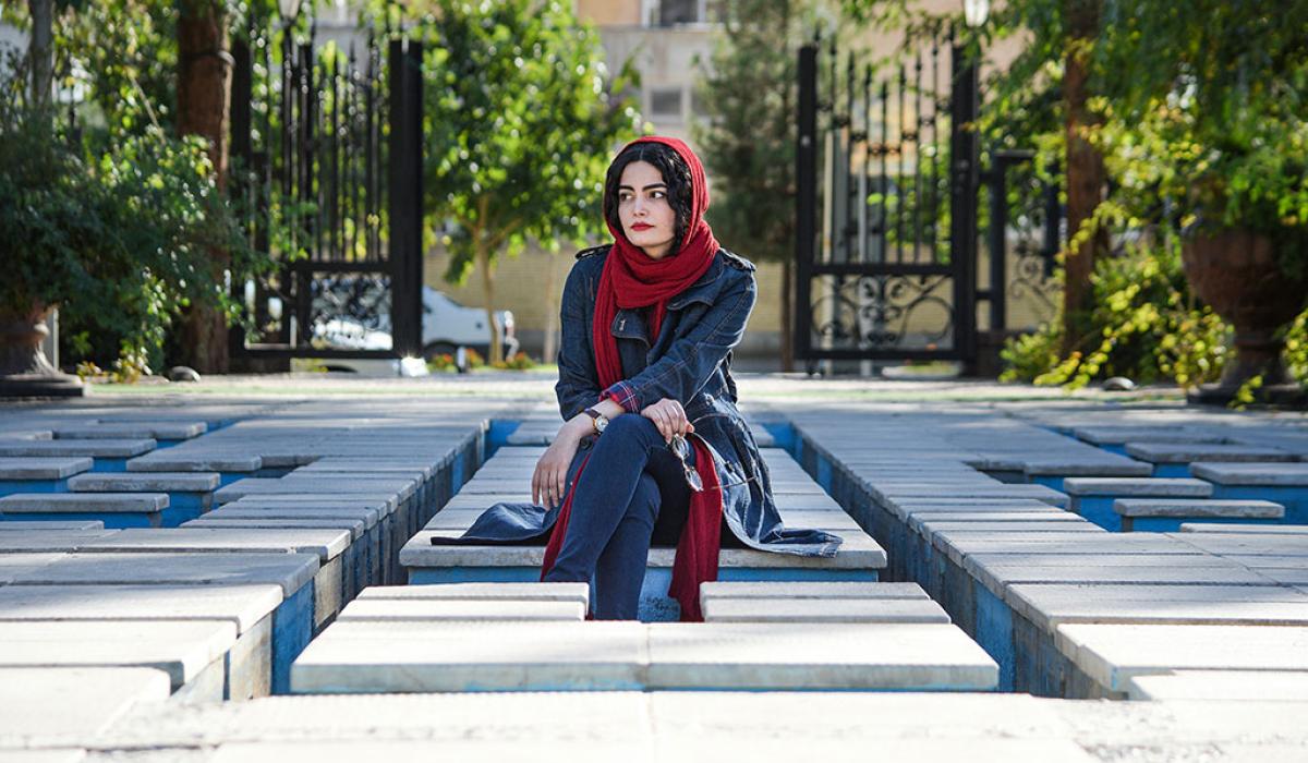 A woman wearing a headscarf sits, looking pensive, amid a grid of concrete seats.