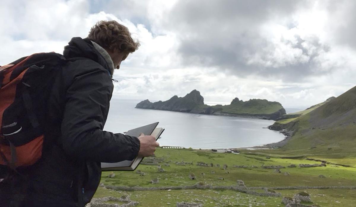 An artist holds a sketchbook while standing overlooking a deserted village by a bay, sided by jagged cliffs.