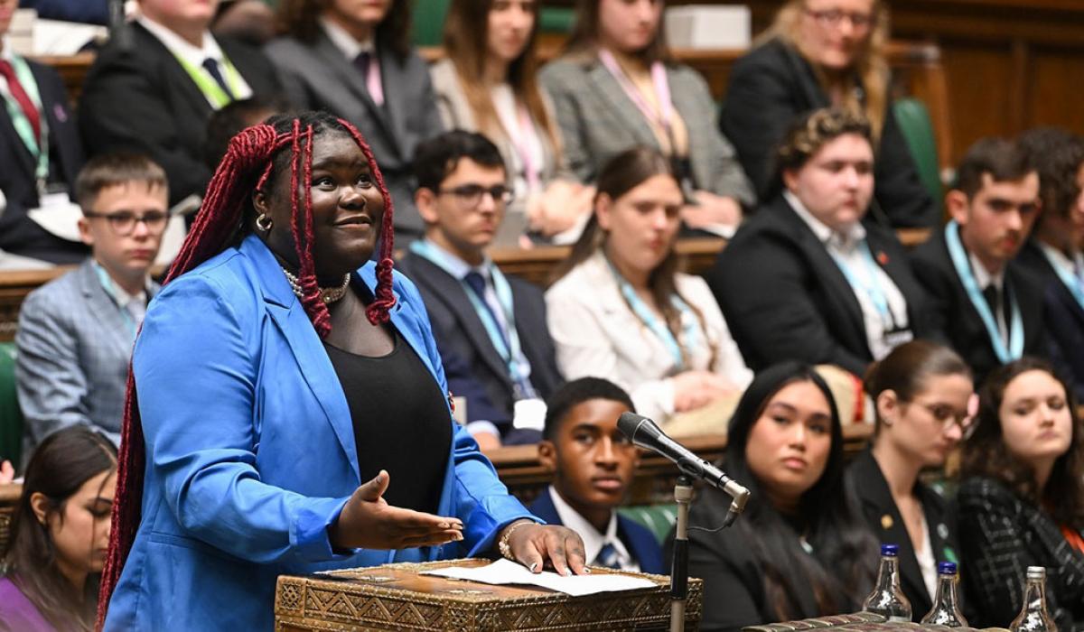 A young woman in a blue suit stands at a wooden box in a parliamentary debating chamber looking upward while speaking. 