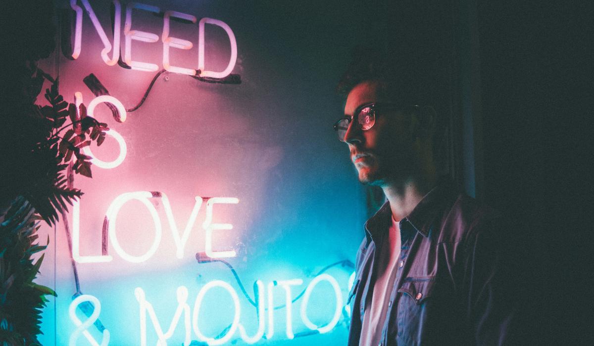 A man stands and looks at a neon sign reading 'need love and... '