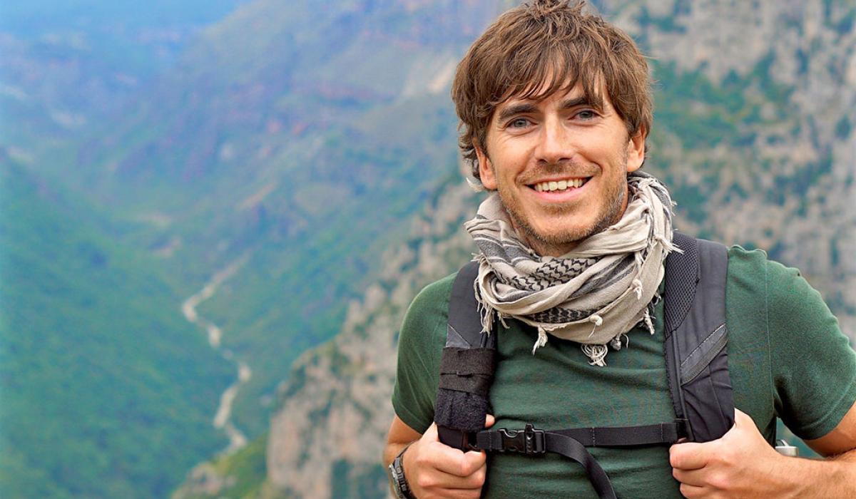 An enthusiastic hiker stands in front of a view down a valley, smiling and holding his backpack straps.