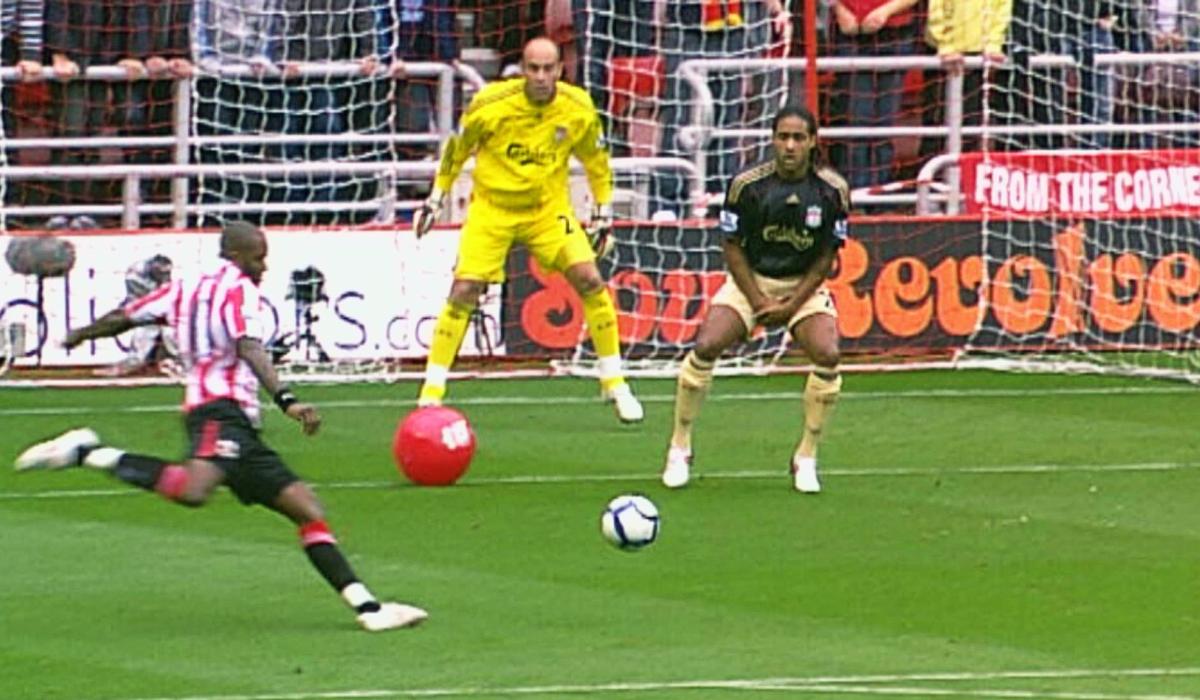 A striker is about kick a football towards a goal, a red beach ball sits between him and the goalkeeper.