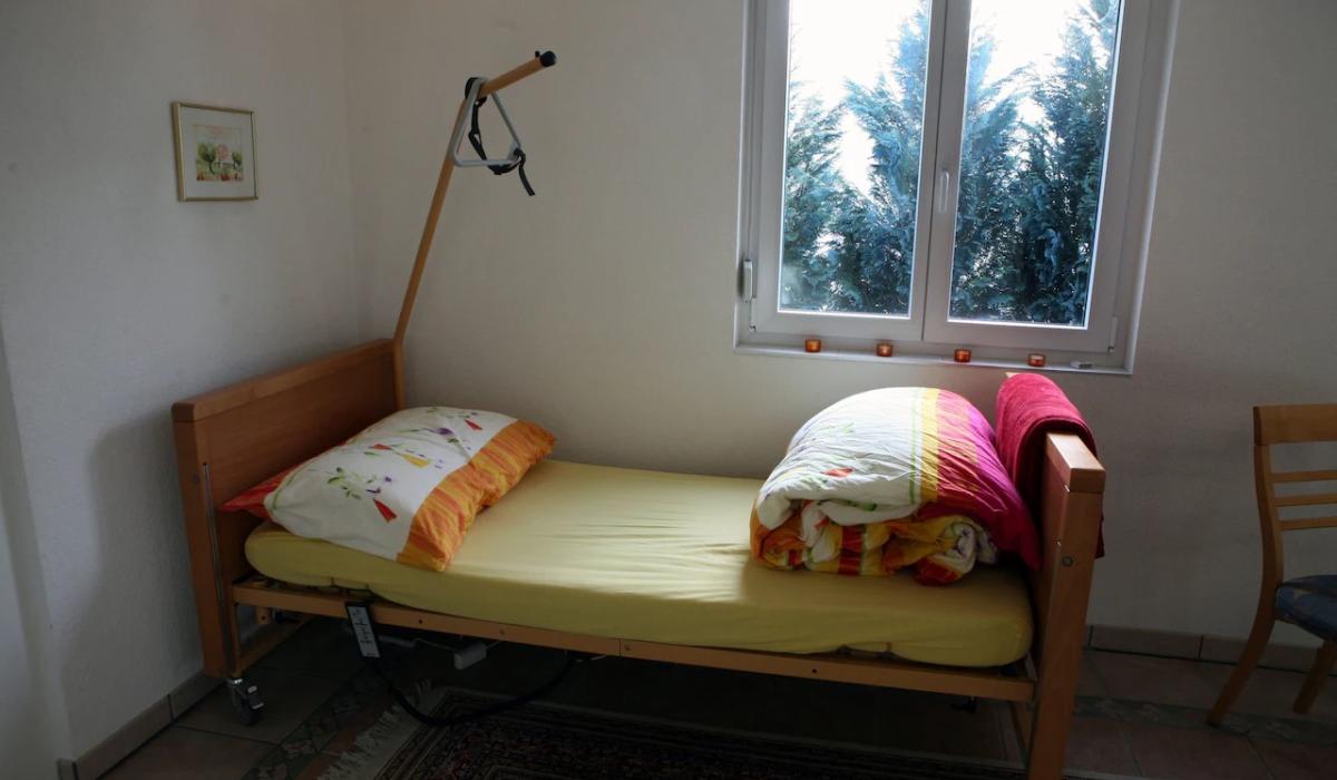 A single bed, wiith an unmade colourful duvet stands in the corner of a room. A hoist reaches over it from the corner. 