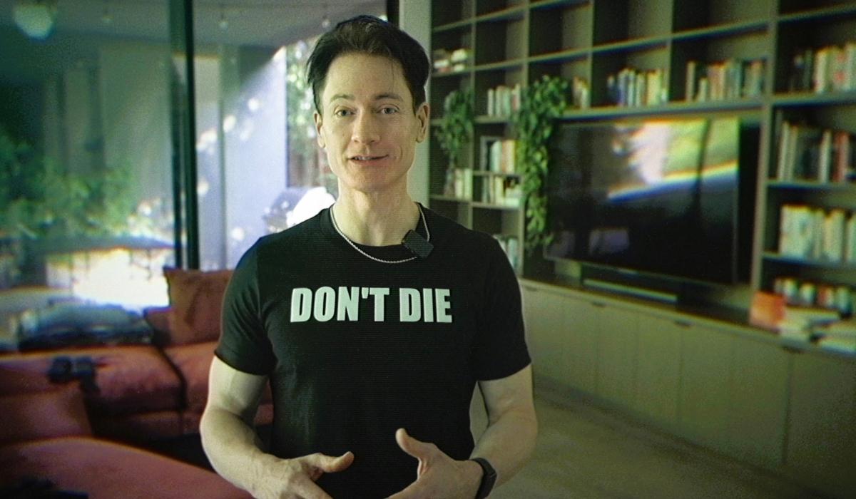 A man stands in his home wearing a black t-shirt that reads 'DON'T DIE'.