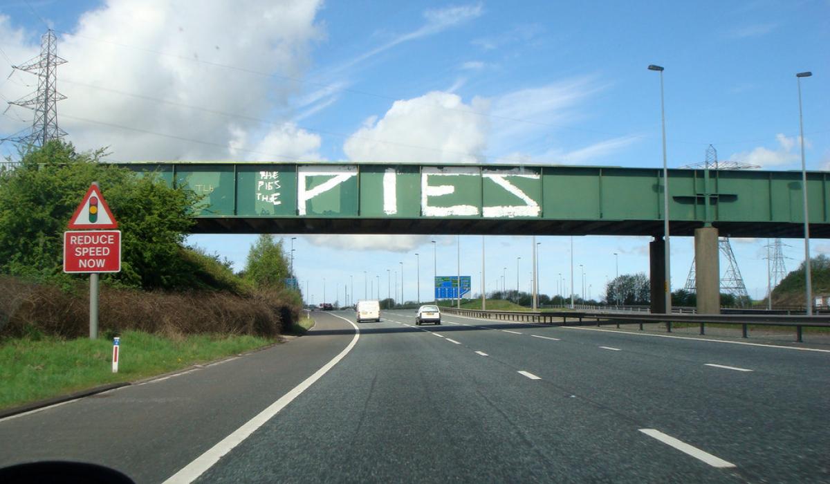 A green bridge spans a motorway, on its side is graffiti that reads 'PIES'