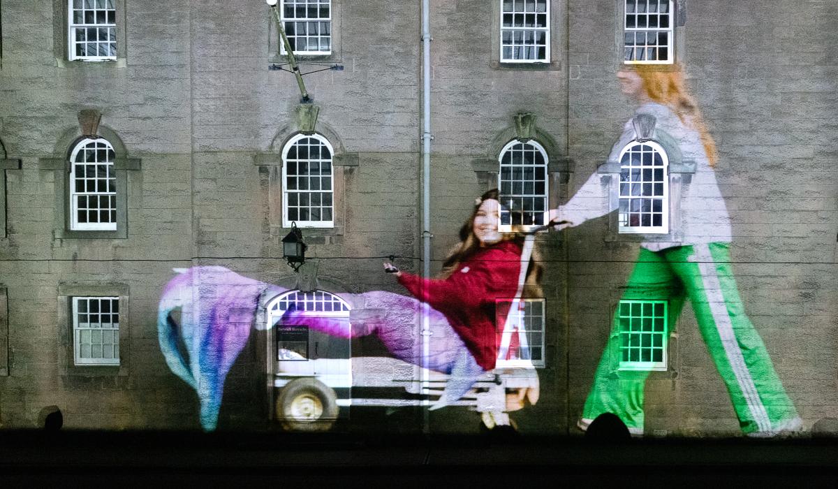 A projected image of a person dressed as a mermaid being pushed on a trolley falls on the wall and windows of an old barracks.