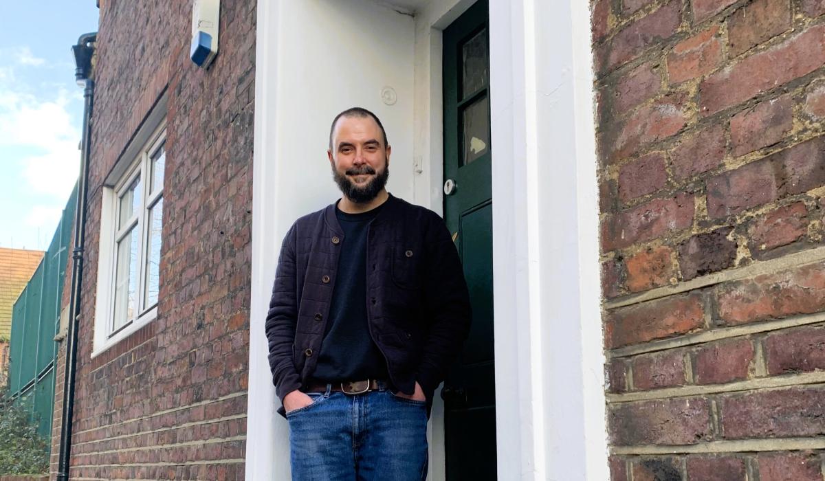 A man stands in a relaxed way on the doorstep of a brick building.