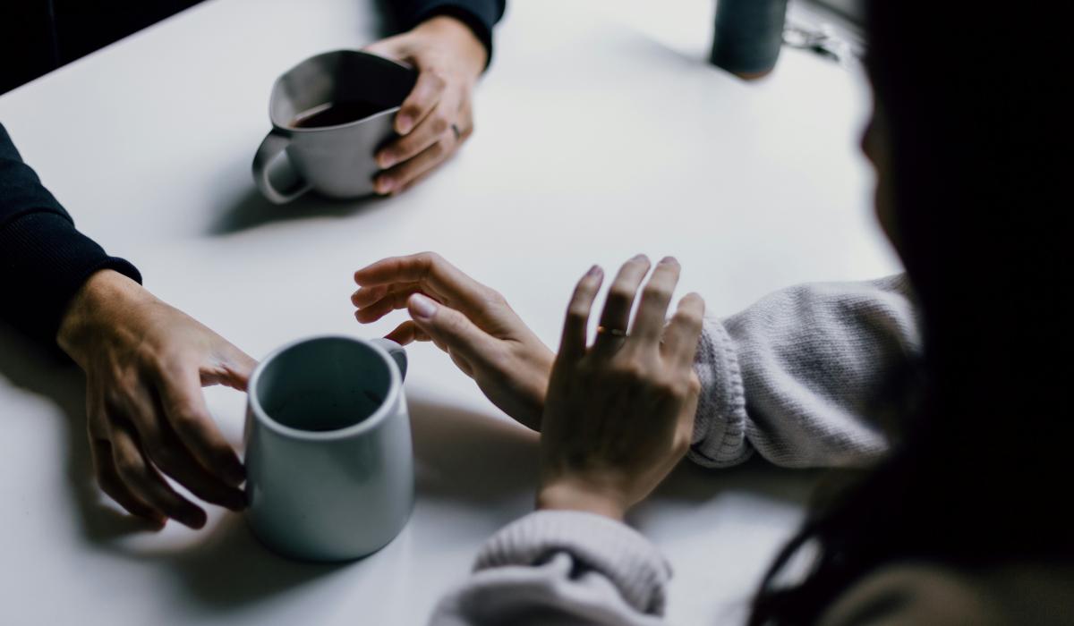 A cup of coffee is offered across a table to a hand that is hesitant.