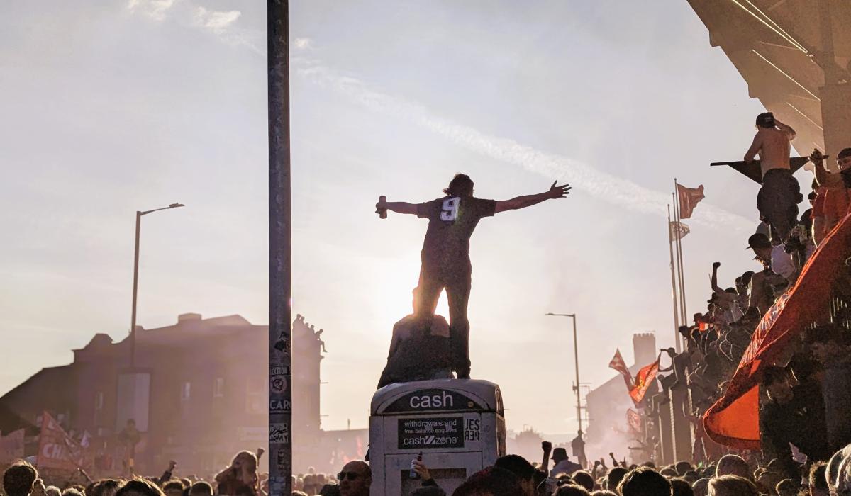 Amid celebrating football fans, one stands on top of a kiosk with outstretched arms.