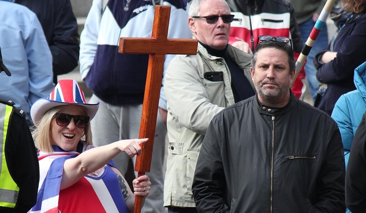 A protester wearing a Union Jack flag and hat and holding a cross, points while a man looks on.