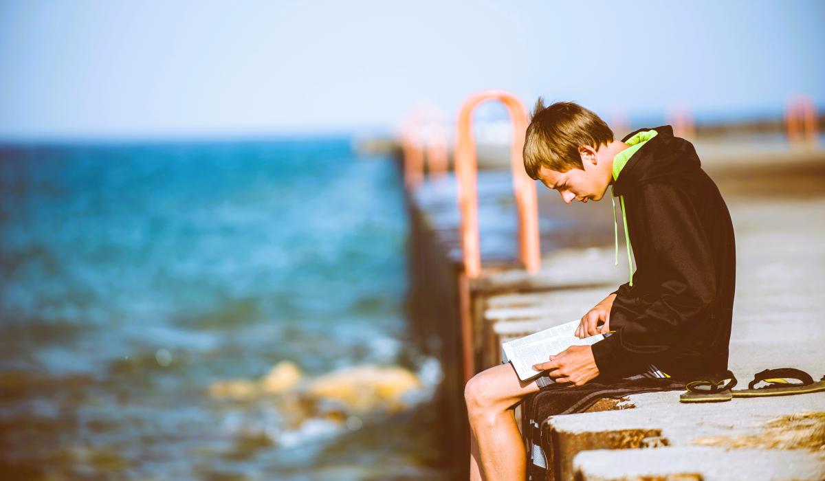 A man sits on a pier intently reading a book on his lap.
