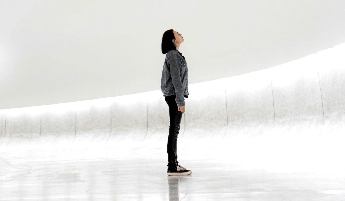 A woman stands against a sparse white background looking up.