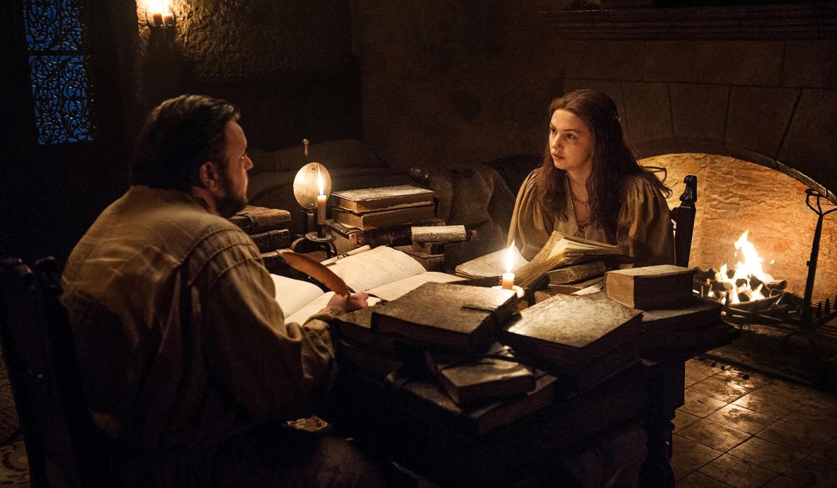 Two people sit at a table strewen with old books lit by candle light.