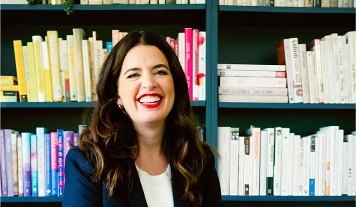 A woman sits and smiles in front of her bookcase.